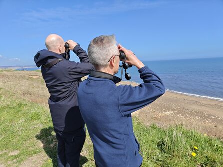 Two people stand on a grassy cliff overlooking a sandy beach and calm blue sea, each holding binoculars and looking out over the water on a bright, clear day.