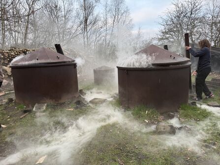Three large metal charcoal kilns stand on grassy ground, each releasing thick white smoke, with a person inspecting one of them beside stacked logs and leafless trees.