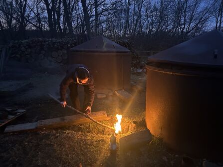 A person stands at dusk tending a small fire with a long stick beside two large metal charcoal kilns, with a tall stack of cut logs and bare trees silhouetted in the background.