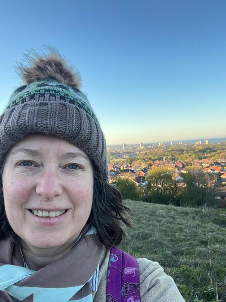 A woman wearing a knitted hat with a pom‑pom and a patterned scarf smiles for a selfie on a grassy hill overlooking a town. The landscape below shows rooftops, trees, and distant tall buildings under a clear blue sky, suggesting a crisp, sunny day.