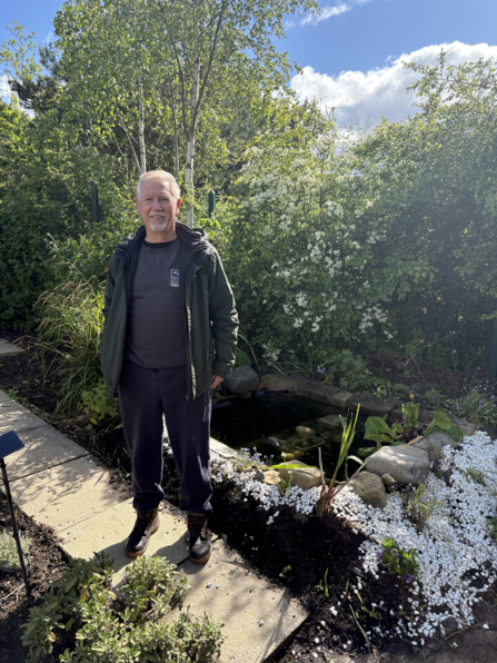 A smiling man wearing a dark jacket, black trousers, and sturdy boots stands on a garden path beside a small pond surrounded by rocks, white gravel, and flowering plants. Behind him, tall birch trees and dense green shrubs with white blossoms fill the background under a bright blue sky with scattered clouds.