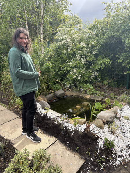  A person wearing a green jumper and black trousers stands smiling beside a small garden pond surrounded by rocks, white gravel, and flowering plants. Behind them, tall birch trees and dense green shrubs with white blossoms create a lush backdrop under a partly cloudy sky.