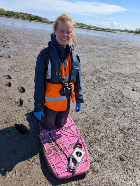 A smiling person wearing a navy waterproof jacket, orange high‑visibility vest, and life jacket stands on a muddy estuary or tidal flat. They have binoculars around their neck and blue gloves on their hands. Beneath them is a pink board with a metal grid and a surveying instrument resting on top, used for fieldwork. The background shows wide mudflats, shallow water, and distant greenery under a bright, partly cloudy sky.