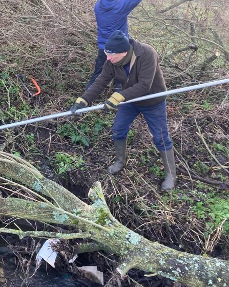 Two people work outdoors beside a small stream clearing fallen branches. The person in the foreground wears a brown jacket, blue trousers, gloves, and rubber boots, using a long metal pole to move a moss‑covered tree trunk lying across the water. Another person in a blue jacket stands behind, handling branches among leafless shrubs. The ground is muddy and covered with winter vegetation.