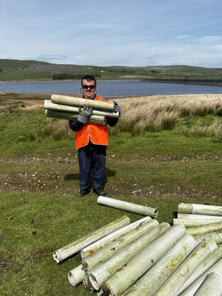 A man wearing a bright orange high‑visibility vest, dark trousers, gloves, and sunglasses stands on grassy terrain beside a lake and rolling hills. He’s smiling while carrying several pale green plastic tubes, with a large pile of similar tubes lying on the ground nearby. The scene shows outdoor conservation or maintenance work under a clear blue sky.