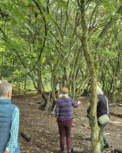 Three people walk through a dense woodland area, following a rope strung between moss-covered trees. The ground is earthy and scattered with fallen branches, while sunlight filters through the green canopy above. The group appears to be taking part in a guided outdoor or sensory activity among the trees.