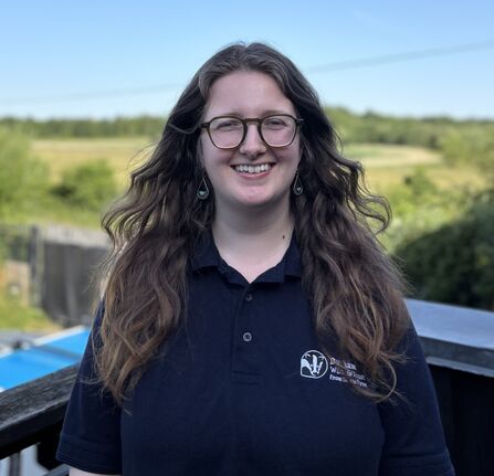A woman with long wavy brown hair and glasses stands smiling outdoors on a sunny day. She wears teardrop earrings and a navy polo shirt with the Durham Wildlife Trust logo and text reading “From the Tees to the Tyne.” Behind her is a green rural landscape with trees and fields under a bright blue sky.