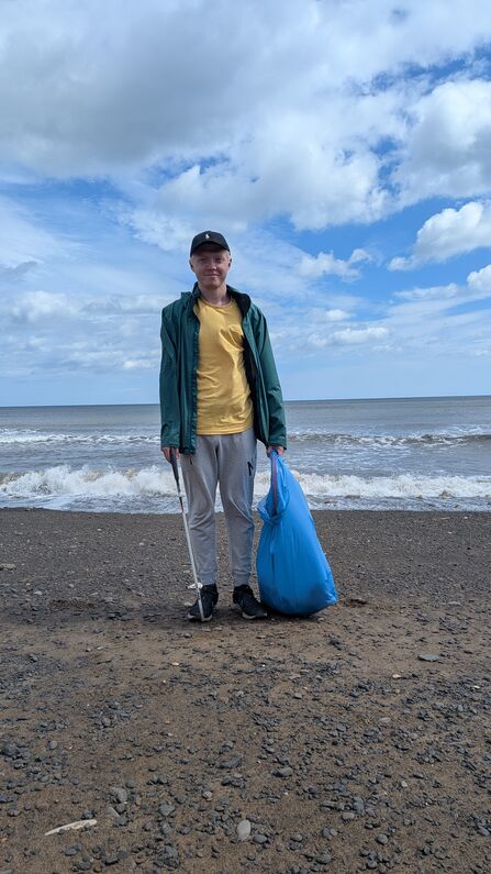 A person stands on a pebbly beach under a partly cloudy sky, holding a blue rubbish bag and a litter picker. They wear a dark green jacket, yellow T‑shirt, grey joggers, black trainers, and a black cap. Waves roll gently behind them, suggesting a coastal clean‑up activity.