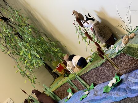 A handcrafted riverside diorama featuring knitted animals and scenery. A badger in a brown jacket and red tie stands by a blue yarn river holding a small creature and leafy branch. Nearby, other knitted animals—like a mole and mouse—sit among green grass, cattails, and white flowers. A tall artificial tree with green leaves and a black bird completes the indoor display.