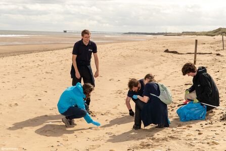 Four volunteers crouch on a sandy beach collecting litter, while another stands nearby observing. They wear gloves and outdoor clothing, using blue bags for rubbish. The sea and a long pier stretch into the distance under a cloudy sky, highlighting the group’s coastal cleanup efforts.
