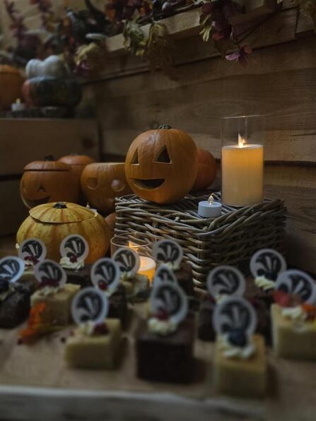 A cosy autumn display featuring carved pumpkins with smiling faces, flickering candles, and an assortment of small decorated cakes. The pumpkins sit on wicker baskets and wooden crates, surrounded by dried leaves and seasonal decorations. The warm candlelight and rustic setting create a festive Halloween atmosphere.