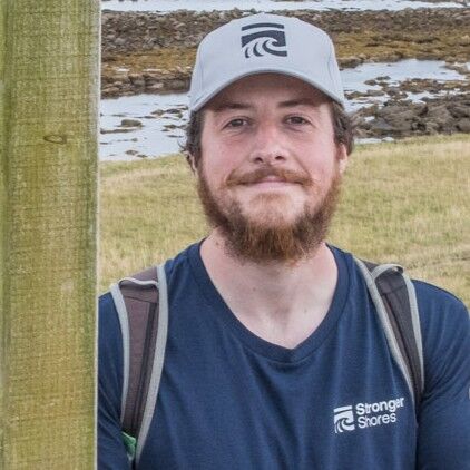 A man with a beard wearing a grey “Stronger Shores” cap and navy T‑shirt stands outdoors near a rocky shoreline and grassy coast, smiling at the camera.