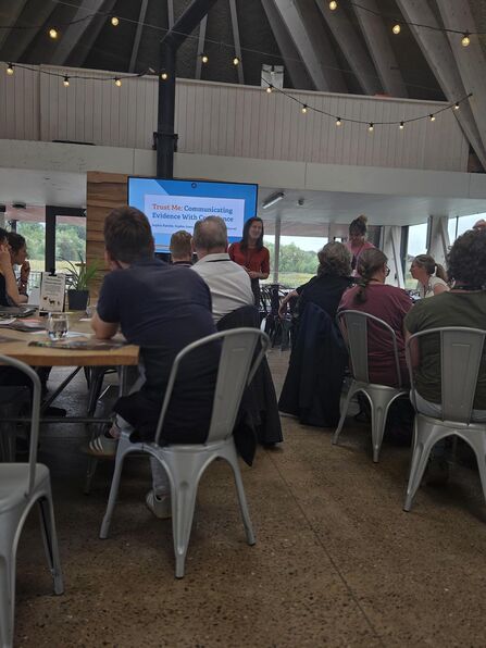 A group of people sit around tables in a bright, modern meeting space with high wooden beams and string lights overhead. At the front, a presenter stands beside a large screen displaying a slide titled “Trust Me: Communicating Evidence With Confidence.” Attendees listen attentively, creating a collaborative workshop atmosphere.
