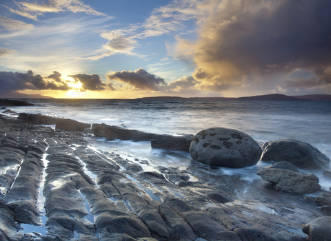 A coastal landscape, with the sea gently lapping at smooth rocks as the sun sets behind scattered clouds