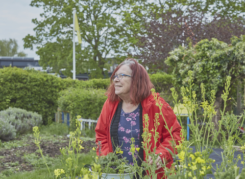 A woman stands by a vegetable patch in a community garden, surrounded by greenery, holding a bowl of freshly collected vegetables.