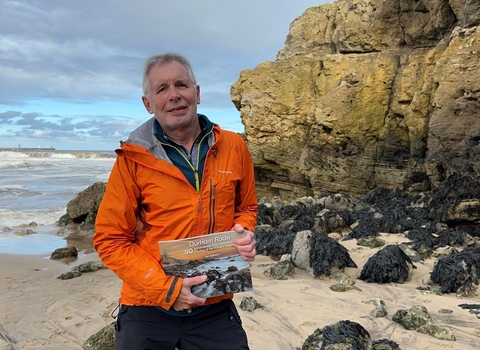 Man standing on beach in front of rock formation holding book