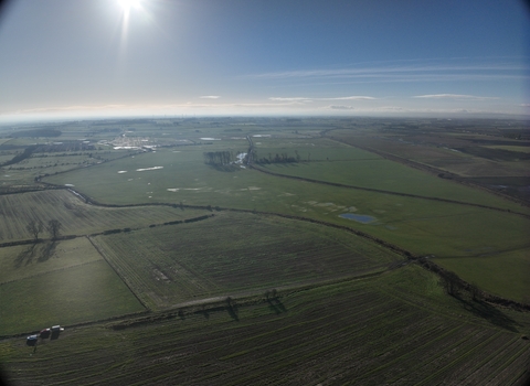 Aerial view over fields with river running through, winter sunlight in distance.