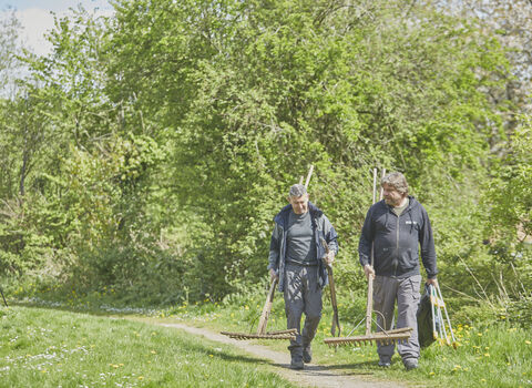 two people walking along pathway carrying gardening tools with grass on either side and trees in background