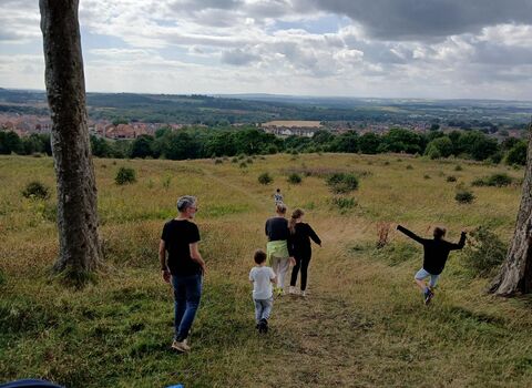 A group of adults and children walk down a grassy hill toward a distant village, surrounded by countryside.