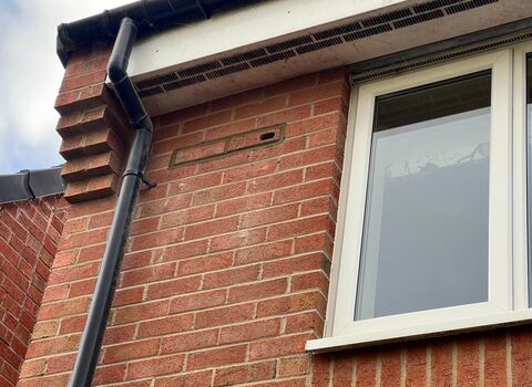 Close-up of a red-brick house wall with a white-framed window, a black downpipe, and a small vent near the roofline beneath a white fascia and black gutter.